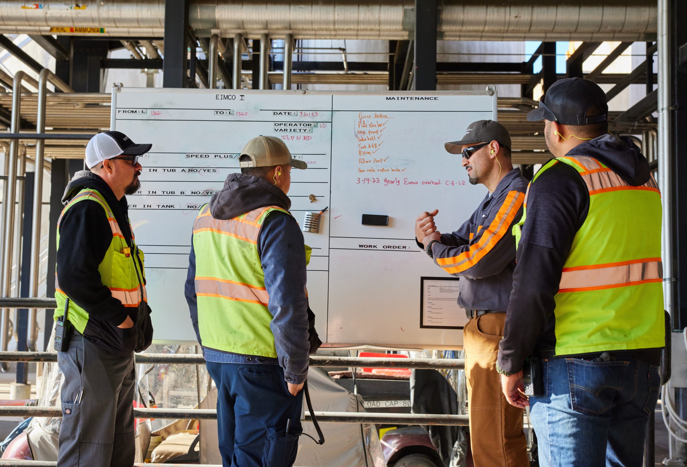 A group of men in construction attire brainstorming around a whiteboard in an open warehouse.