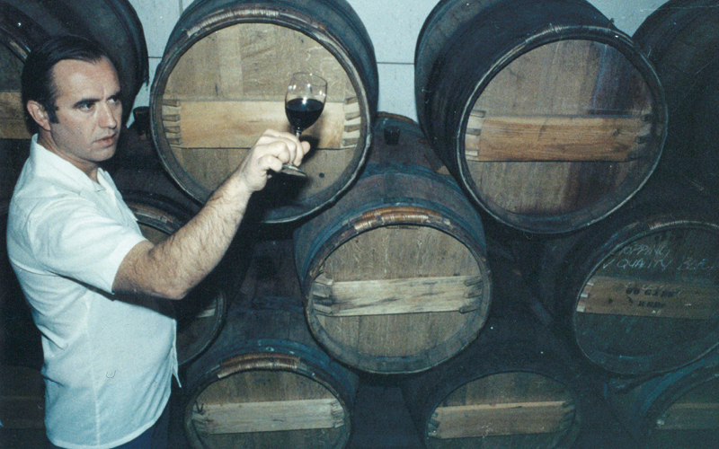 Color image of Bob Trinchero inspecting a glass of red wine in a barrel room.